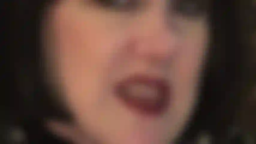 Face closeup in black bob wig, teasing with red lipstick lips and wet tongue as well as teeth and leather gloved hands which would enjoy holding on to your balls.